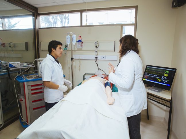 Dos personas enseñando en una sala tipo laboratorio con un muñeco de pruebas.